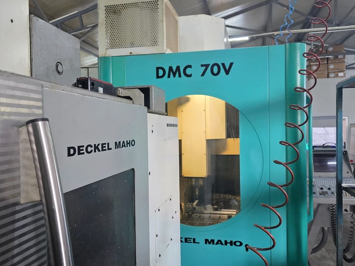 Vertikalni CNC obradni centar DECKEL MAHO DMC 70 V u hali; kroz okno vrata vidi se vreteno iznad stola. Levo u prvom planu je susedna DECKEL MAHO mašina.
