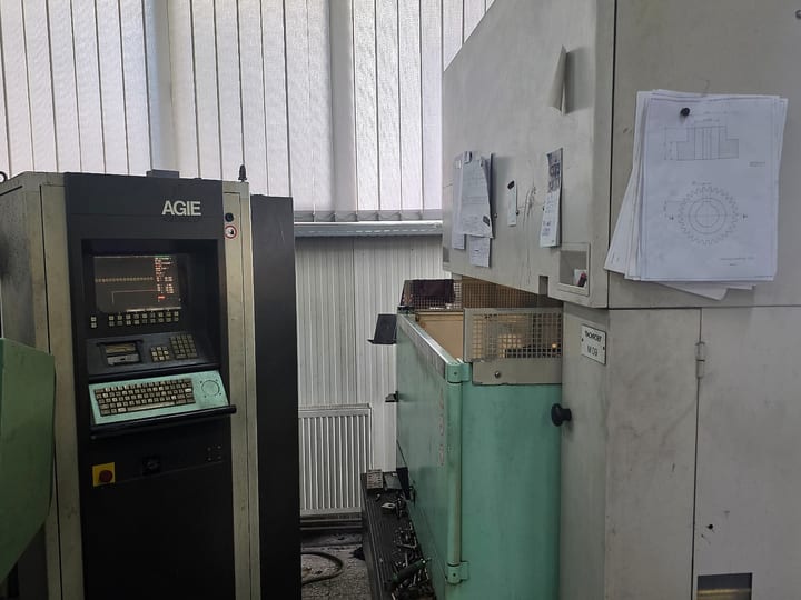 Upravljački ormar AGIE (AGIEMATIC CF) sa ekranom i tastaturom, pored radnog suda erozimata; na kućištu mašine okačeni su tehnički crteži.