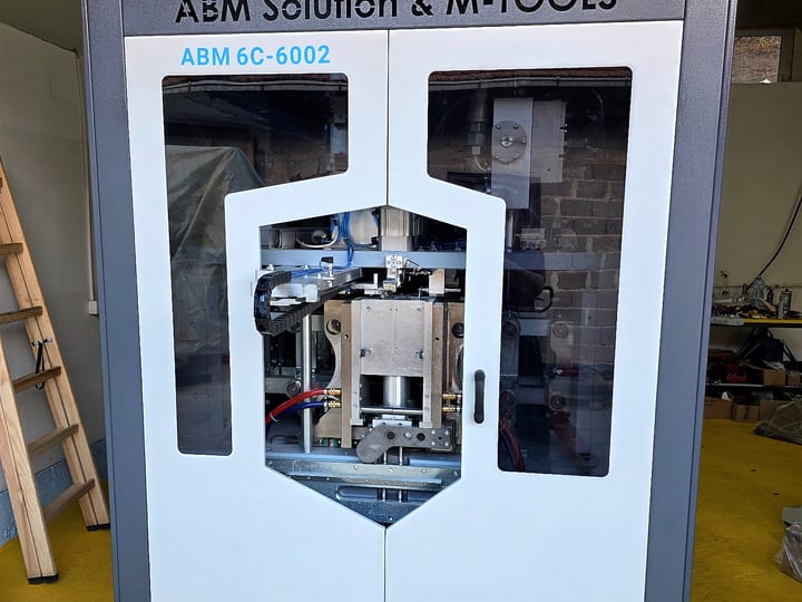 Automatska PET duvaljka ABM 6C-6002 u belom kućištu sa natpisom ABM Solution & M-TOOLS; mehanizam se vidi kroz staklena vrata.
